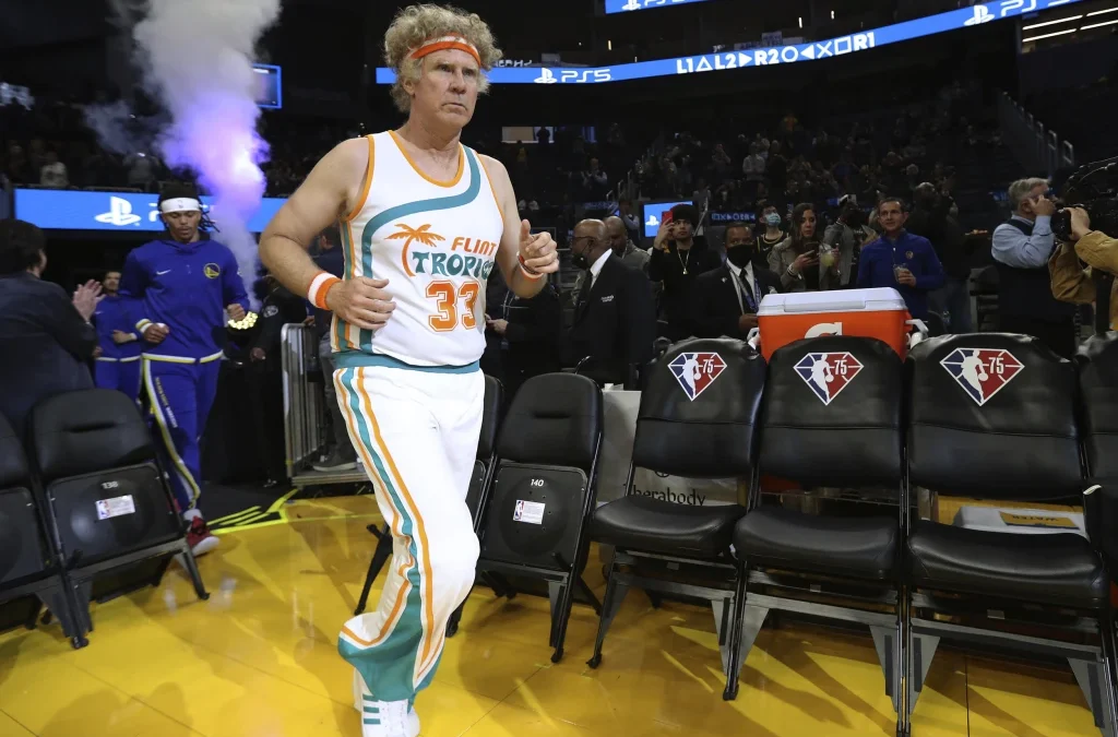 Will Ferrell in a white, teal, and orange ‘Flint Tropics’ semi-pro basketball uniform jogging onto a court